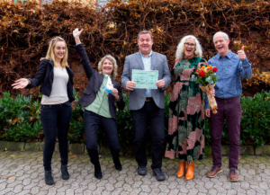 Fem glade personer står foran en hekk og viser frem blomster, en skulptur og en gavesjekk